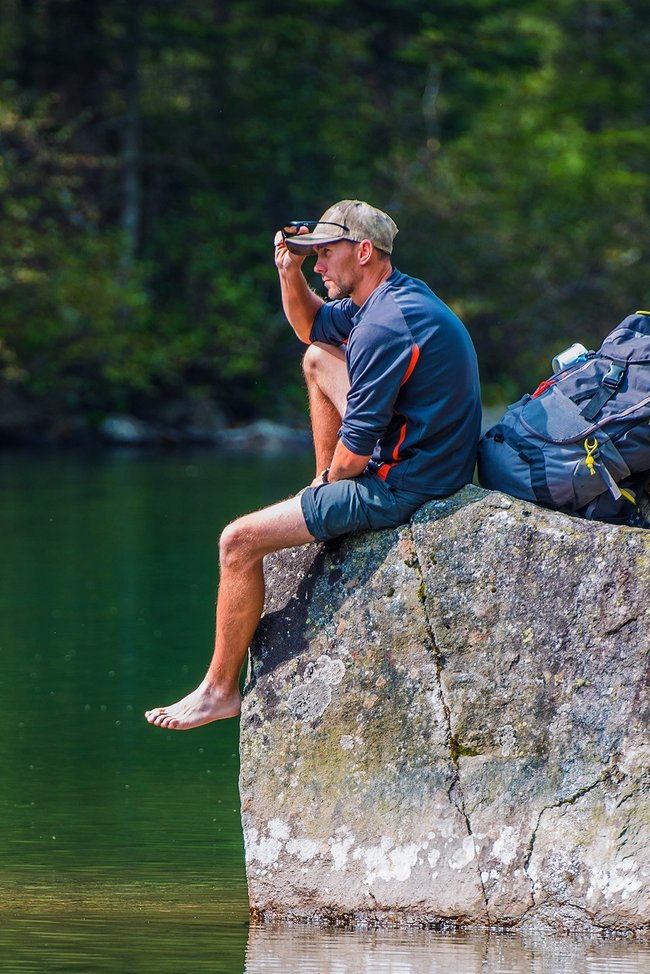 Wanderer sitzt barfuß auf einem Stein am Ufer eines Sees